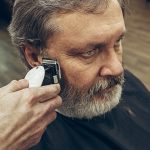 Close-up side view portrait of handsome senior bearded caucasian man getting beard grooming in modern barbershop. Hairdresser serving client, making beard haircut using shaving machine. Barber shop concept.