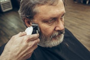 Close-up side view portrait of handsome senior bearded caucasian man getting beard grooming in modern barbershop. Hairdresser serving client, making beard haircut using shaving machine. Barber shop concept.